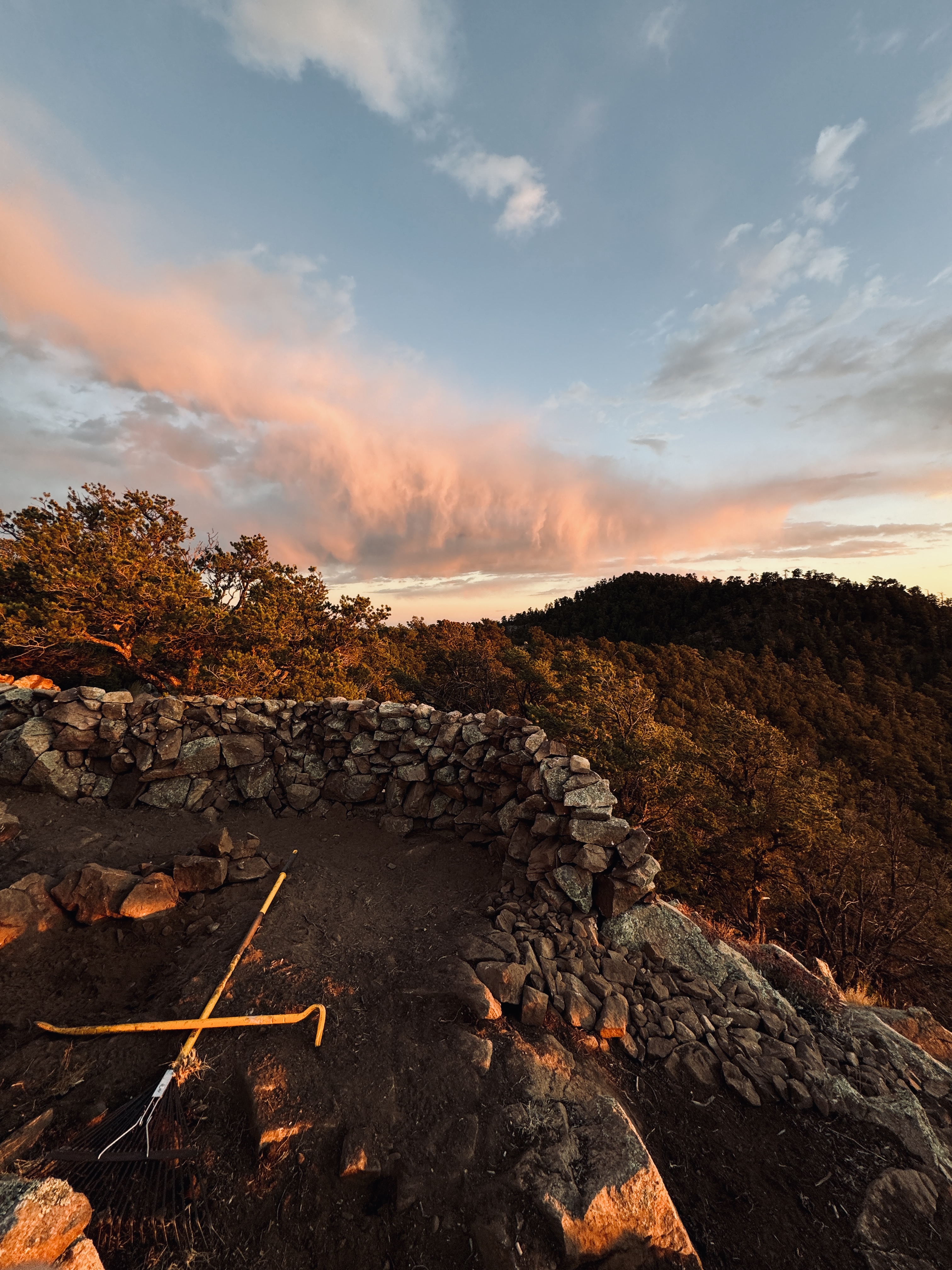 Dry-stacked stone structure on ridgeline at sunset — pickaxe, clouds, piñon trees